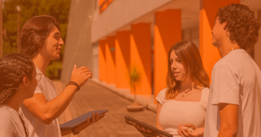 Un grupo de cuatro jóvenes de la Universidad Anáhuac Veracruz se encuentra conversando animadamente en un entorno exterior, con un edificio de fondo que presenta colores naranjas
