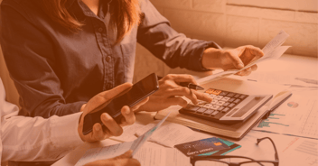 Un grupo de personas está revisando documentos financieros sobre una mesa, ocupándose activamente en tareas de cálculo. Una mano sostiene un teléfono móvil, mientras que otra usa una calculadora, rodeada de hojas con gráficos y estados financieros.