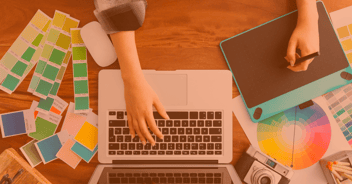 Un escritorio creativo con un laptop abierto y herramientas de diseño. Una mano está en el teclado, mientras que la otra sostiene un marcador sobre una tableta gráfica. Alrededor, hay muestras de colores y una rueda de colores, junto con una cámara vintage