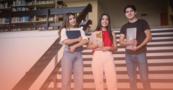 Tres estudiantes universitarios, dos mujeres y un hombre, sonriendo y sosteniendo cuadernos y laptops, posan de pie en las escaleras de una biblioteca con estanterías llenas de libros al fondo. La imagen representa el ambiente estudiantil, el aprendizaje de idiomas y las habilidades interculturales en la educación superior.