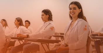 Estudiantes con bata blanca en un aula, una joven al frente sonríe a cámara; todos llevan el logo de la Universidad Anáhuac.