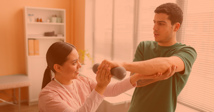 Una mujer guía a un hombre mientras realiza ejercicios con un par de mancuernas en un entorno de rehabilitación. La mujer sonríe y sostiene una de las mancuernas, mientras el hombre se concentra en el ejercicio. La habitación tiene luz natural y estanterías al fondo