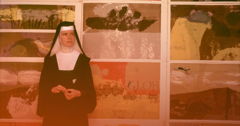 Corita Kent, vestida como monja, de pie frente a una pared cubierta con sus obras de arte coloridas y abstractas.
