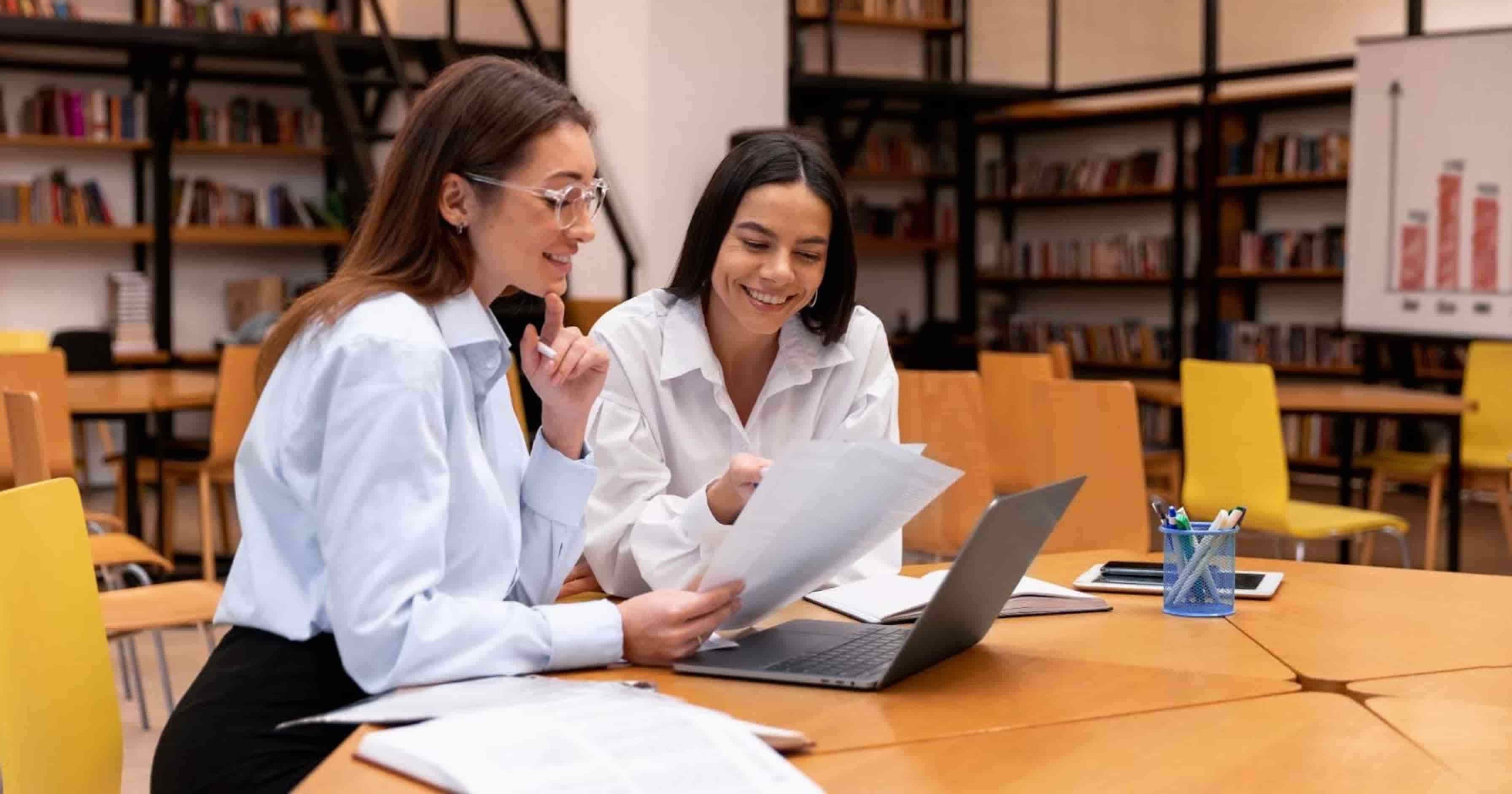 Dos mujeres profesionales revisando documentos y gráficos en una biblioteca, analizando oportunidades después de concluir un diplomado.